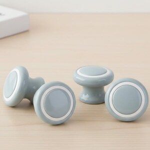 Puede incluir: Cuatro pomos de cerámica azul claro con un anillo blanco. Los pomos tienen forma redondeada y están colocados sobre una superficie de madera clara. Estos pomos decorativos son ideales para renovar muebles.