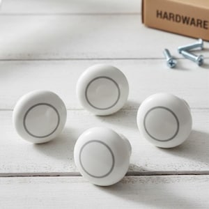May include: Four white ceramic cabinet knobs with a grey circular design. The knobs are round and sit on a white wooden surface. A box labelled "HARDWARE" is in the background.