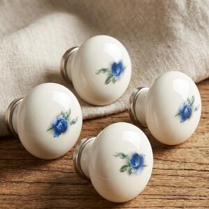 Puede incluir: Cuatro pomos de armario de cerámica blanca con base plateada y diseño de rosa azul. Los pomos están colocados sobre una superficie de madera con un paño de color neutro en el fondo. Herrajes decorativos.