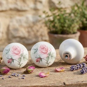 Könnte beinhalten: Drei weiße Keramik-Schrankknöpfe mit einem rosa Rosenmuster. Die Knöpfe sind rund mit flachem Boden und einer Metallschraube. Getrocknete Rosenknospen und Lavendelzweige sind auf einer Holzoberfläche verstreut.