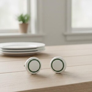 May include: Two white cabinet knobs with a green ring design. The knobs are round and sit on a light-coloured wooden surface. A stack of white plates and a small potted plant are visible in the background.