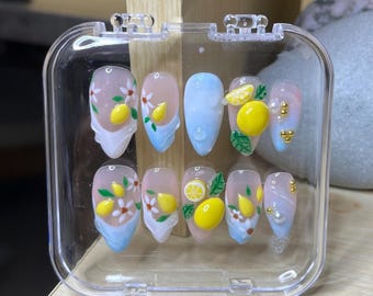 Puede incluir: Un juego de diez uñas artificiales en una caja de plástico transparente. Las uñas presentan una variedad de diseños, incluyendo limones, flores blancas y efectos de ombre azul y blanco. Algunas uñas tienen detalles dorados y perlas.