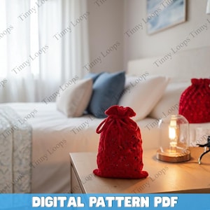 Może przedstawiać: Dwie czerwone, szydełkowe torby ze sznurkiem stoją na drewnianym stoliku nocnym. Torby są teksturowane i mają marszczony wierzch. Tekst "DIGITAL PATTERN PDF" znajduje się na dole obrazu. W tle znajduje się łóżko z poduszkami.