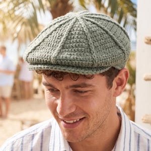 Pode incluir: Boné newsboy de crochê verde sálvia. O boné tem um design texturizado e canelado e uma aba curta. A imagem mostra o boné na cabeça de uma pessoa, com uma cena de praia ao fundo.