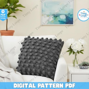 May include: A dark gray crocheted pillow with a textured, bobble stitch design rests on a white sofa. The image includes a blanket, a potted plant, and a side table with flowers. The text "DIGITAL PATTERN PDF" is at the bottom.