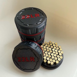 May include: Black cylindrical container with red accents and the text "22LR" in red. The container is partially open, revealing a collection of small, brass-colored objects. The container is sitting on a white surface.