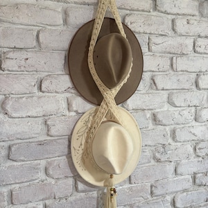 May include: A cream-coloured macrame hat holder with two hats suspended against a white brick wall. The top hat is brown, the bottom hat cream with embroidered leaves. The macrame has a wooden ring at the top.