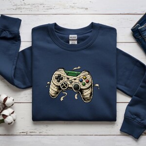 May include: Navy blue sweatshirt featuring a mummy-themed video game controller graphic. The controller has beige bandages, colorful buttons, and a green top. The sweatshirt is made of a soft material.