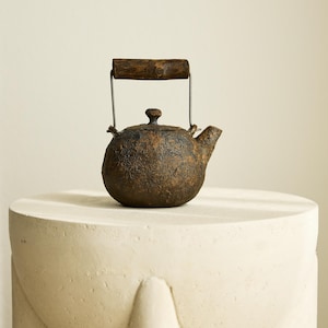 Op de afbeelding: Een kleine, ronde theepot met een getextureerde, donkerbruine buitenkant en een houten handvat. De theepot heeft een tuit en een kleine knop op het deksel. Het handvat is bevestigd met metaaldraad. De theepot staat op een witte, ronde sokkel.