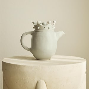 Può includere: Una teiera in ceramica grigio chiaro con un coperchio a forma di corona. La teiera ha un corpo arrotondato, un manico curvo e un beccuccio. È posizionata su un piedistallo chiaro e strutturato. Il coperchio a corona presenta piccoli dettagli arrotondati.
