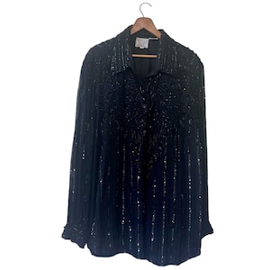May include: Black button-down shirt with all-over sequin embellishments. The shirt is hanging on a wooden hanger. The sequins create a textured, shimmering effect. The shirt has a collared neckline and long sleeves.
