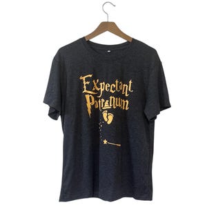 May include: Dark gray t-shirt with the words "Expectant Patronum" in gold lettering, along with a baby footprint and a wand. The shirt is hanging on a wooden hanger.