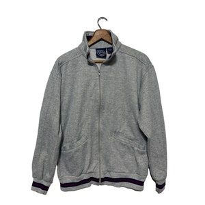 May include: A gray zippered sweatshirt with a ribbed collar and cuffs. The sweatshirt has a small logo on the left chest and a pocket on the front. The cuffs and waistband have a red and blue stripe.