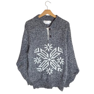 May include: A gray and white snowflake pattern sweater with a button-up collar. The sweater is hanging on a wooden hanger against a white background. The snowflake design is centered on the front of the sweater.