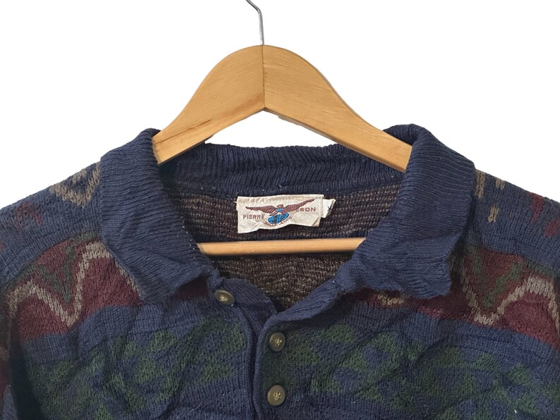 May include: A blue and brown patterned knit sweater with a collared neckline and a button closure. The sweater has a label that reads "Pierre Cardin".