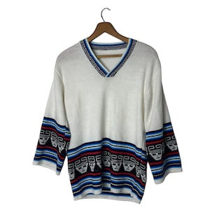 May include: White sweater with a V-neck and a geometric pattern on the sleeves. The pattern is in shades of blue, red, and black.