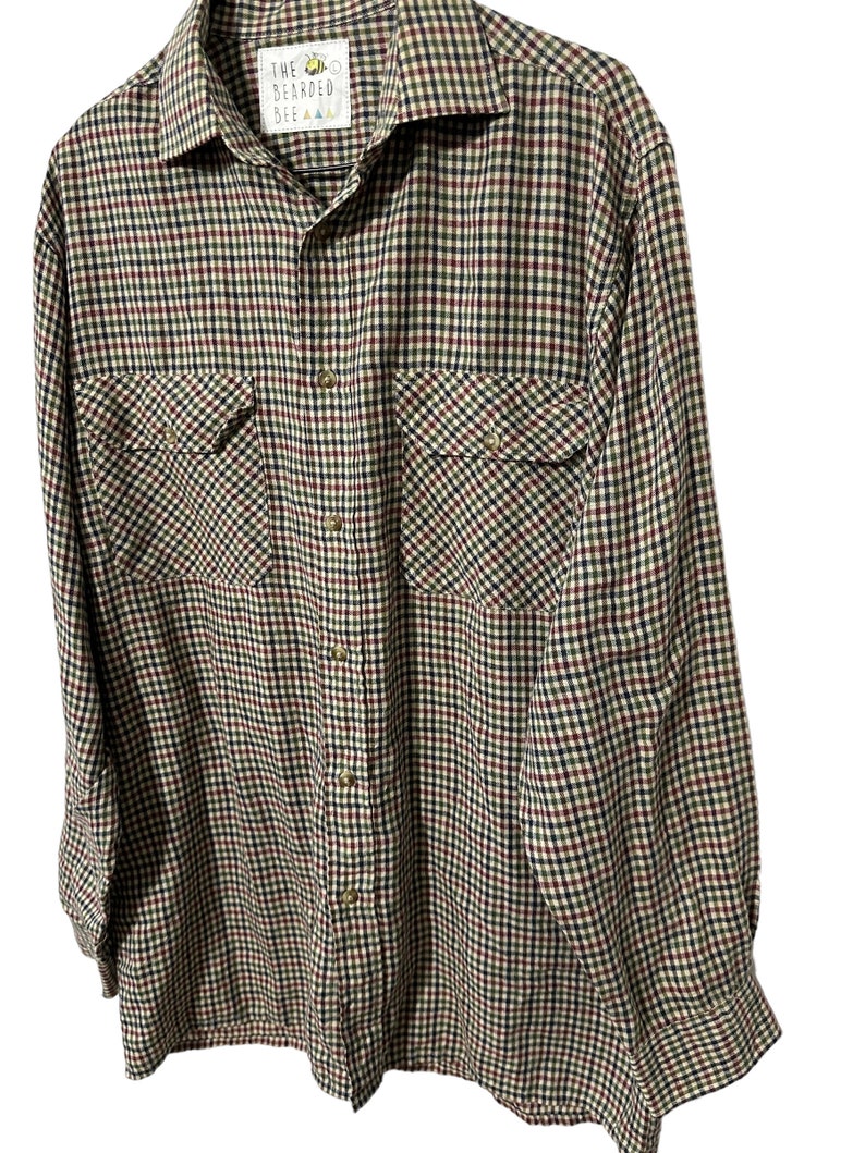 May include: A long-sleeved plaid shirt in a brown, green, red, and black checkered pattern. The shirt has a button-up front and two chest pockets. The label on the shirt reads "The Bearded Bee".