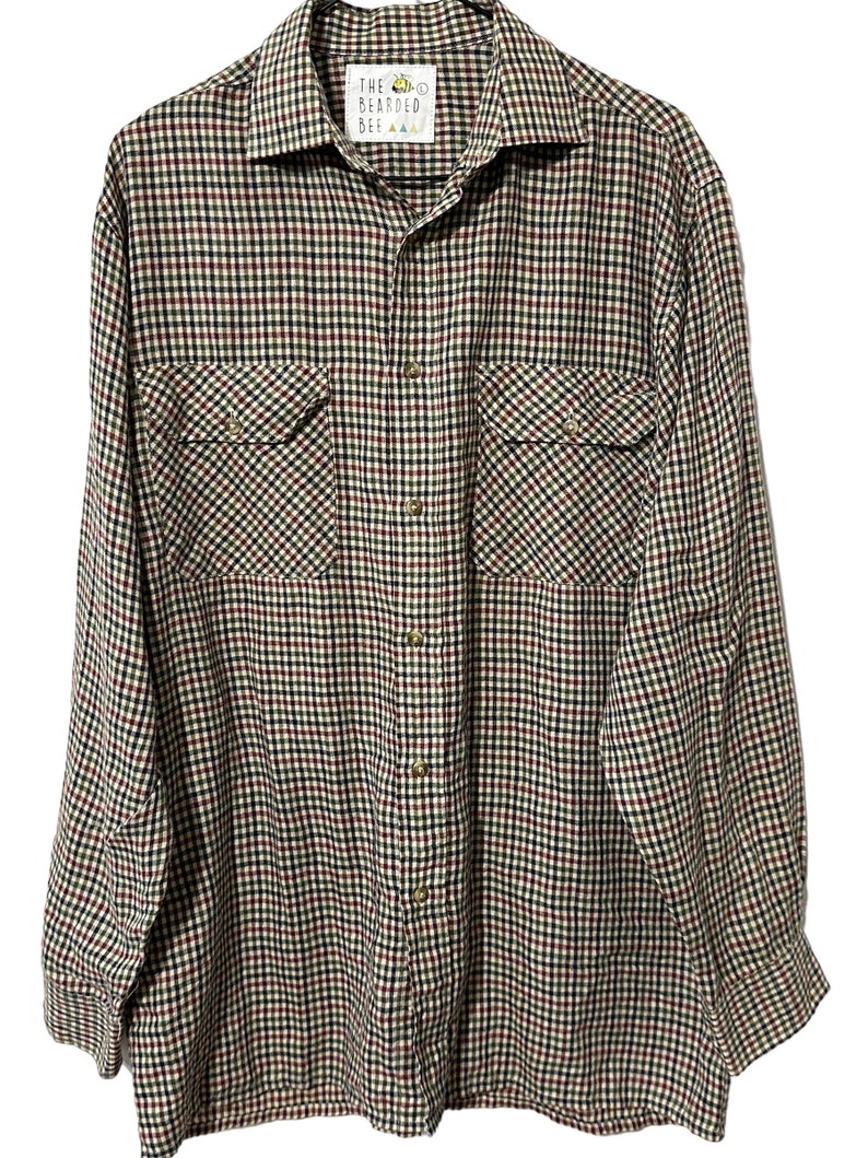 May include: A long-sleeved plaid shirt with a button-up front and two chest pockets. The shirt is in a black, red, and green plaid pattern. The label on the shirt reads "The Bearded Bee".