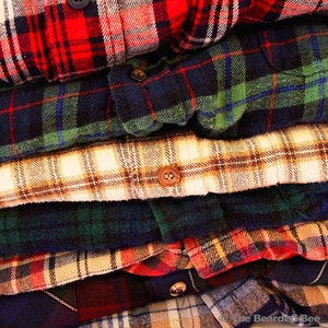May include: A stack of plaid flannel shirts in various colours, including red, blue, green, and tan. The shirts are folded neatly and have a rustic, vintage aesthetic.