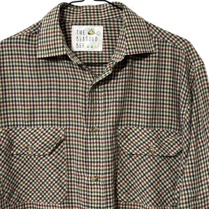 May include: A long-sleeved plaid shirt with a button-up front and two chest pockets. The shirt is in a beige, red, black, and green plaid pattern. The label on the shirt reads "The Bearded Bee".