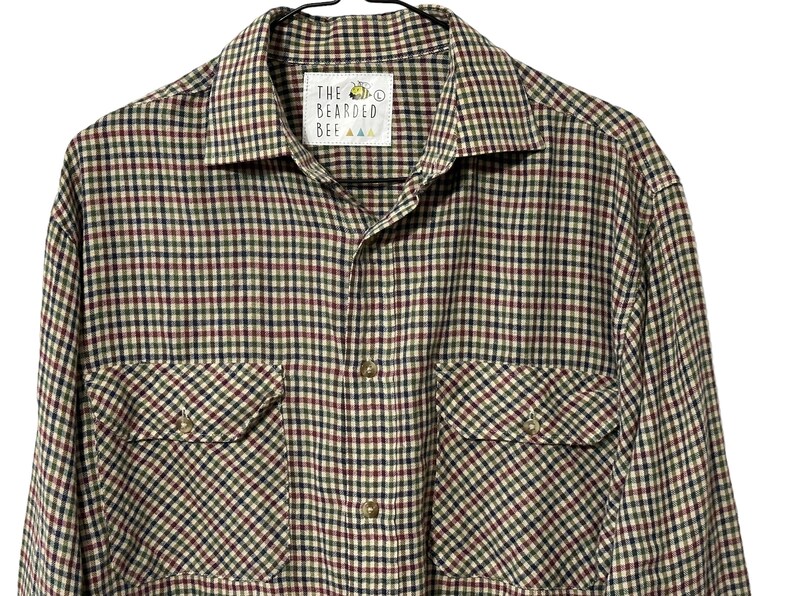 May include: A long-sleeved plaid shirt with a button-up front and two chest pockets. The shirt is in a beige, red, black, and green plaid pattern. The label on the shirt reads "The Bearded Bee".
