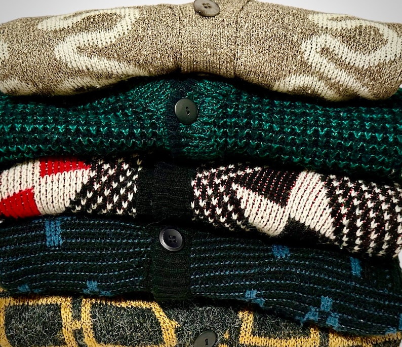 May include: A stack of folded cardigans in various colors and patterns. The top cardigan is beige with a white zigzag design. Below it is a dark green cardigan, followed by a red, black, and white patterned cardigan. The stack includes other patterned cardigans.
