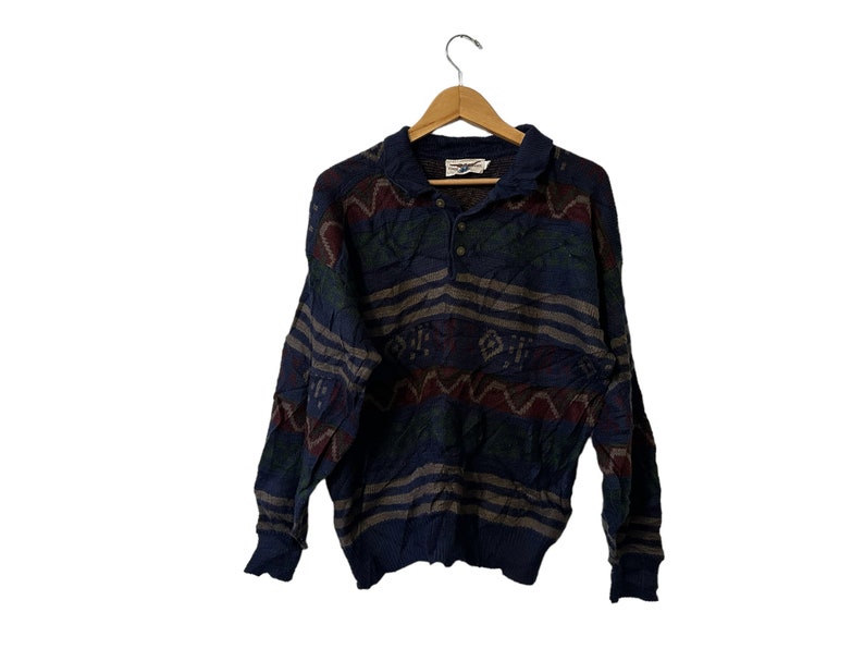 May include: A blue, green, and red patterned long-sleeve sweater with a button-up collar. The sweater has a woven design and is hanging on a wooden hanger.