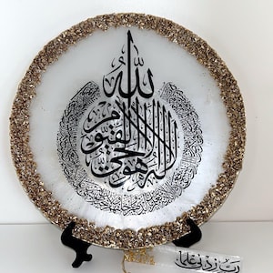 Peut inclure: Assiette décorative blanche avec une bordure en paillettes dorées et une calligraphie arabe noire. L'assiette est présentée sur un support noir. Un marque-page transparent avec un pompon doré et du texte arabe est également visible.