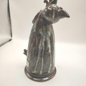 May include: A handcrafted ceramic rooster figurine. The sculpture features a brown glaze with white and green accents, and a detailed design. The rooster has a long neck, a curved beak, and a decorative crest. The base is round and sturdy.