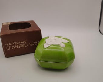 Op de afbeelding: Een felgroene keramieken doos met een witte, gebeeldhouwde roos op het deksel. Daarnaast staat een bruine kartonnen doos met de tekst FINE CERAMIC COVERED BOX in het wit op de voorzijde gedrukt. De keramieken doos heeft een gladde, glanzende afwerking.