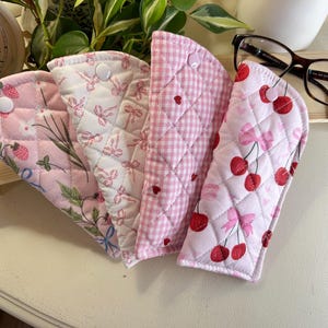 Könnte beinhalten: Vier gesteppte Brillenetuis in rosa und weißen Mustern. Designs umfassen Erdbeeren, Schleifen, Vichy-Karo und Kirschen. Jedes Etui hat einen Knopfverschluss. Eine Brille liegt auf einem kleinen Holzregal.