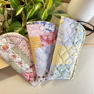 May include: Three quilted eyeglass cases in patchwork patterns. Each case features a different fabric design, including floral, geometric, and solid colors. The cases are secured with a white button closure. A pair of eyeglasses rests on a wooden shelf.