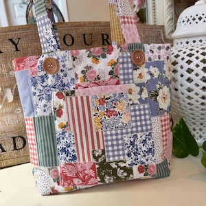 Puede incluir: Un bolso tote patchwork con una variedad de telas estampadas florales y otros diseños en tonos azules, rosas, rojos y verdes. El bolso tiene dos cierres de botón de madera y asas de tela a juego.