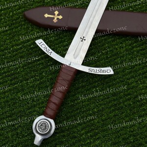 May include: A medieval-style sword with a silver blade and a brown leather-wrapped handle. The sword features a cross design and the words "CHRISTUS" and "VINCIT" on the blade. A brown leather sheath with a gold cross is visible in the background.