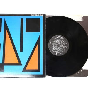 May include: Split Enz "True Colours" vinyl record album. The album cover has a blue background with orange and black geometric shapes. The record is black, and the sleeve shows a black and white photo of a person.