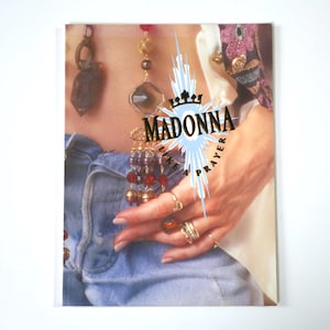Könnte beinhalten: Ein Magazin-Cover mit dem Text "MADONNA Like a Prayer" in einem Sternmuster. Das Bild zeigt eine Hand mit Ringen, die eine Halskette hält. Die Person trägt eine Jeans.
