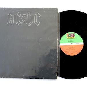 May include: A black vinyl record album titled Back in Black by AC/DC. The square cardboard sleeve features the band name AC/DC in white outline text. The vinyl disc is black with a circular Atlantic label featuring green and orange sections.
