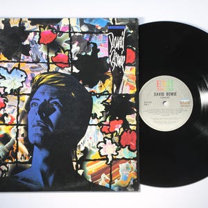 May include: A David Bowie Tonight vinyl record and its square cardboard sleeve. The cover features a blue-toned portrait of David Bowie against a grid background filled with colorful floral and abstract patterns. The record is black vinyl.