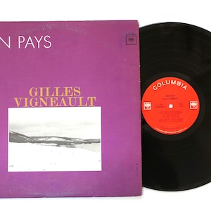May include: A vintage vinyl record album with a purple cover featuring the text "MON PAYS" and "GILLES VIGNEAULT." The album is next to a black vinyl record with a red label that reads "COLUMBIA."