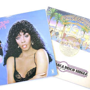 May include: Two vinyl record covers. The left cover features Donna Summer with the text "Donna Summer Bad Girls" and a pink street lamp. The right cover shows a Casablanca Record and Filmworks sleeve with palm trees, camels, and "Casablanca Disco Single".