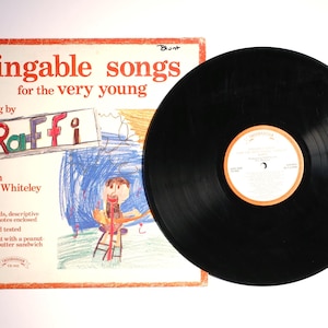May include: A vintage vinyl record and album cover for "Singable Songs for the Very Young" by Raffi. The album cover features colorful childlike drawings and text. The record is black with a white and orange label.