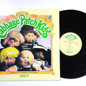 May include: A vintage vinyl record album titled "Cabbage Patch Kids" featuring illustrations of Cabbage Patch Kids dolls. The album cover is light yellow with the title in green and the words "Cabbage Patch Dreams". The record is black.