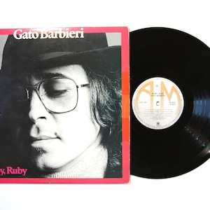 May include: A vinyl record album by Gato Barbieri, titled "Ruby, Ruby." The album cover features a black and white portrait of a man wearing glasses and a hat, set against a red border. The black vinyl record is next to the album cover.