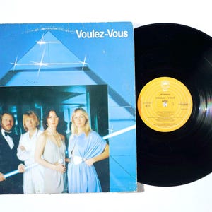 Op de afbeelding: Een vinylplaat en de vierkante kartonnen hoes van het ABBA-album Voulez-Vous. De blauwe hoes toont een foto van de vier bandleden voor een driehoekige structuur met sterachtige lichteffecten. De grammofoonplaat is zwart van kleur.