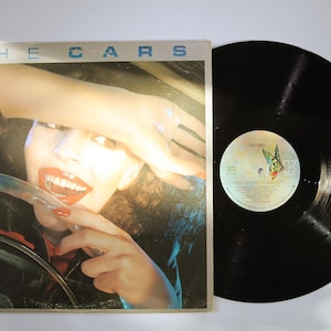 May include: The Cars self-titled vinyl record album with a square cardboard sleeve featuring a photographic portrait of a person with red lipstick and nails behind a steering wheel, alongside a black circular vinyl record with a butterfly label.