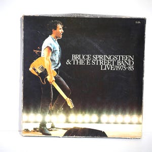 Puede incluir: Portada del álbum "Bruce Springsteen & The E Street Band Live/1975-85". La portada presenta a Bruce Springsteen con una guitarra, con luces de escenario de fondo. El título del álbum está impreso en blanco.