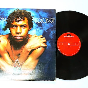 May include: An album cover featuring Edwin Birdsong with the album title "What It Is" in white text. The cover art shows a man with dark hair and a necklace against a blue background. A black vinyl record with a red label is next to the album cover.