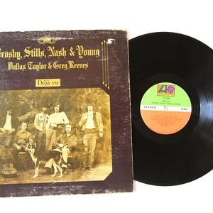 Könnte beinhalten: Vintage Vinyl-Schallplattenalbum mit dem Titel "Déjà vu" von Crosby, Stills, Nash & Young. Das Albumcover zeigt eine Sepia-Fotografie der Band mit einem Hund. Die Schallplatte ist schwarz mit einem farbenfrohen Etikett.