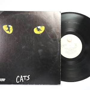 May include: A black vinyl record album cover for the musical Cats, featuring two bright yellow cat eyes with silhouettes inside. The text reads London Original Cast Recording and Cats. A black vinyl record is visible next to the cover.