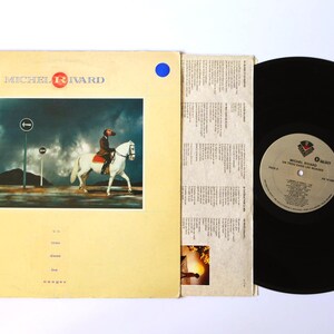 Può includere: Un album in vinile vintage di Michel Rivard. La copertina giallo chiaro presenta un'immagine di una persona a cavallo su un cavallo bianco. Il disco è nero e l'album include un foglio di testi. L'album è in buone condizioni.